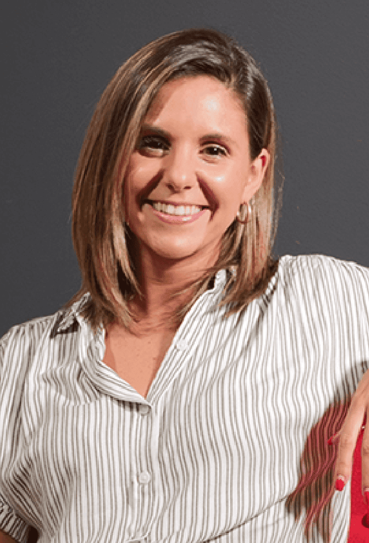 A woman with shoulder-length brown hair, smiling, wearing a white and gray striped shirt, and standing against a gray
