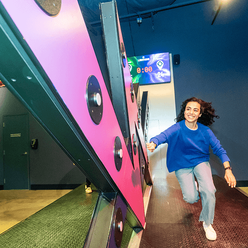 Punch Team Culture Photo 1 A woman in a blue sweater and gray pants is playing a large interactive game with colorful panels and black circular targets.
