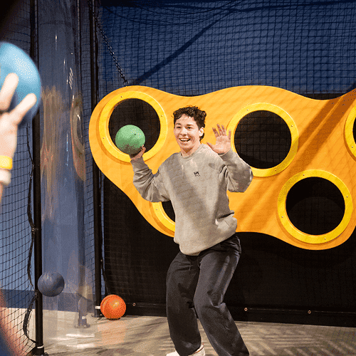 Punch Team Culture Photo 10 A smiling person holding a green ball in an indoor play area with a yellow climbing structure and black netting background.
