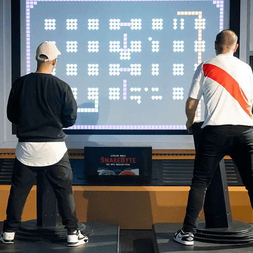 Punch Team Culture Photo 13 Two men stand on pedal-controlled platforms playing a large digital Pac-Man game on a screen.