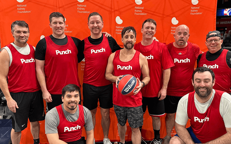 Punch Team Culture Photo-13 A group of nine men in red "Punch" basketball jerseys pose together at a charity event with a bright orange backdrop.