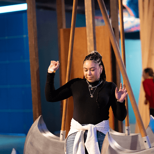 Punch Team Culture Photo 14 A woman with braided hair and layered necklaces stands with her eyes closed, raising her arms in front of a swing set.