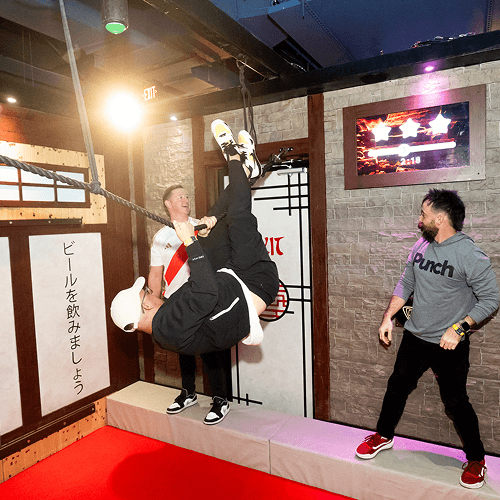 Punch Team Culture Photo 16 A man swings on a rope while another man holds him, with a third person watching in a room with brick walls and a TV screen.