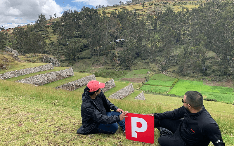 Punch Team Culture Photo-17 Two people sit on a grassy hillside holding a red sign with a white "P" against a backdrop of terraced fields and a forested