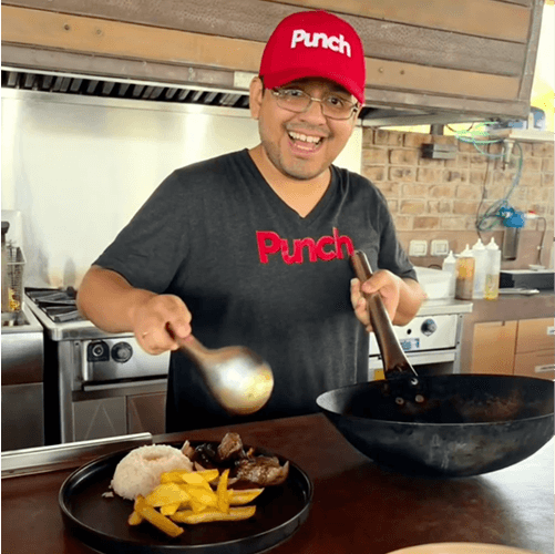 Punch Team Culture Photo-2 A man wearing a red "Punch" hat and black "Punch" shirt cooks food in a kitchen with a wok and a plate of rice, fries, and