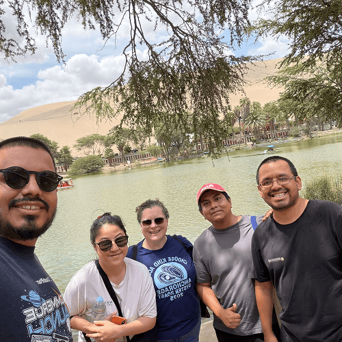 Punch Team Culture Photo 20 Five people stand together by a lake with trees and sand dunes in the background, smiling for a group photo.