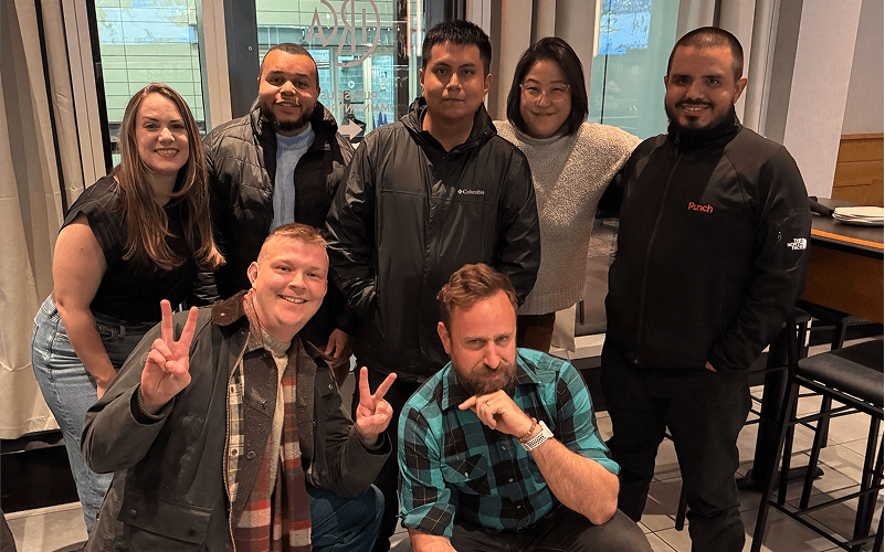 Punch Team Culture Photo-20 Seven people pose together indoors, with two making peace signs and one resting his chin on his hand.