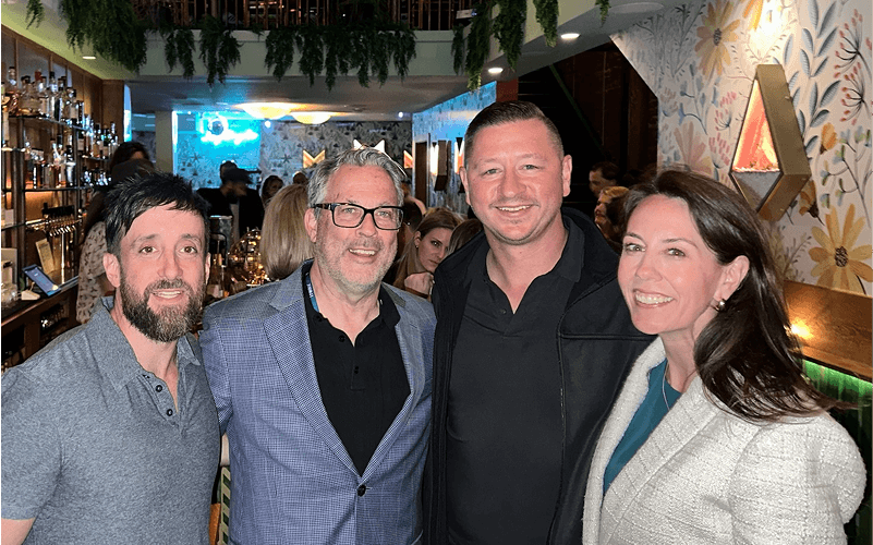 Punch Team Culture Photo-22 Four people stand together smiling in a lively bar or restaurant with floral wallpaper and hanging greenery.