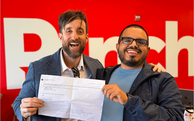 Punch Team Culture Photo-23 Two smiling men hold a document together in front of a red background with the partial word "Pitch" visible.