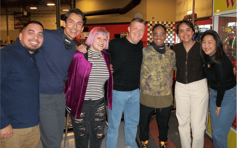 Punch Team Culture Photo-27 Seven people stand together in an arcade, smiling and posing for a group photo.