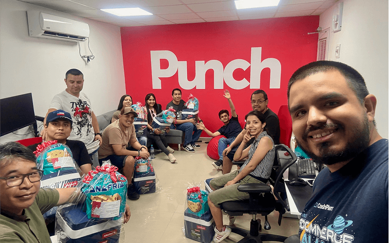 Punch Team Culture Photo-29 A group of people, including children, sitting and standing in an office with a red wall that says "Punch," holding gift