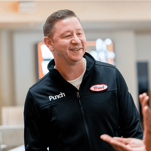 Punch Team Culture Photo 3 A smiling man wearing a black jacket with "Punch" and "Brad" patches greets someone indoors.