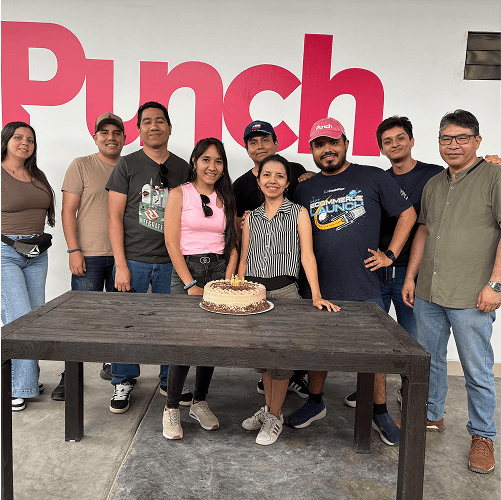 Punch Team Culture Photo-33 Eight people stand behind a table with a birthday cake, celebrating in front of a large "Punch" sign.