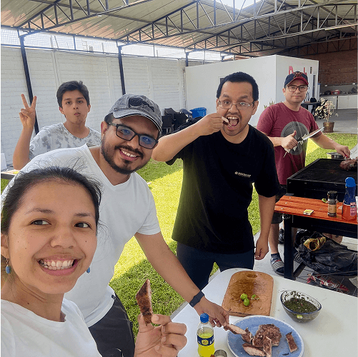 Punch Team Culture Photo-35 Five people are gathered outdoors under a covered area, preparing and enjoying grilled food together.