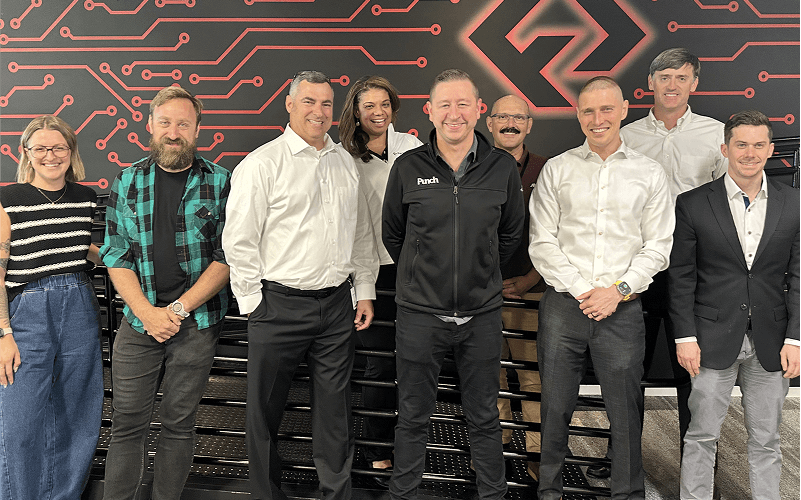 Punch Team Culture Photo-36 A group of nine diverse professionals stands together in front of a high-tech, circuit-themed backdrop with a logo.