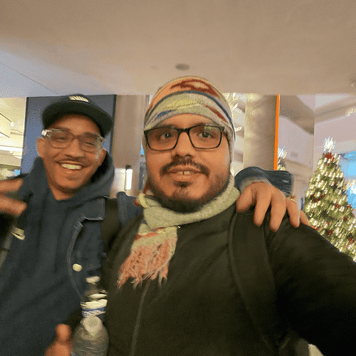 Punch Team Culture Photo 5 Two smiling men, one wearing glasses and a colorful beanie, pose together in front of a decorated Christmas tree.