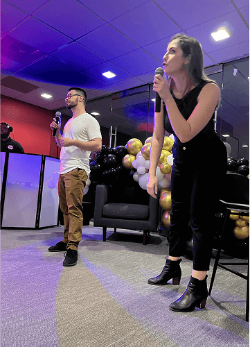 Punch Team Culture Photo-6 A woman and a man are speaking into microphones at an indoor event with black, gold, and white balloons in the background.