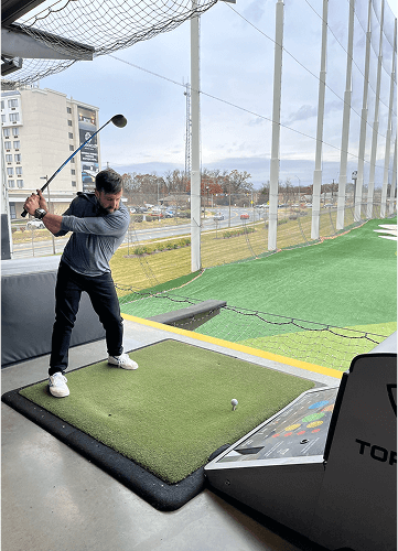 Punch Team Culture Photo-7 A man is practicing golf swings at an indoor driving range with a golf ball on a tee, surrounded by netting and a cityscape