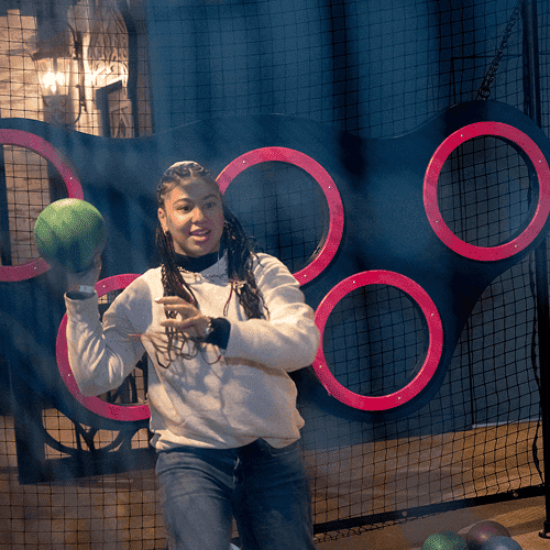 Punch Team Culture Photo 9 A young woman with braided hair holds a green ball at an indoor play area with pink circular frames and a netted background.