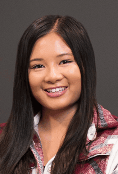A young woman with long, straight black hair smiling against a gray background.
