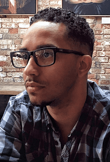 A man with short curly hair, glasses, and a plaid shirt sitting in front of a brick wall.