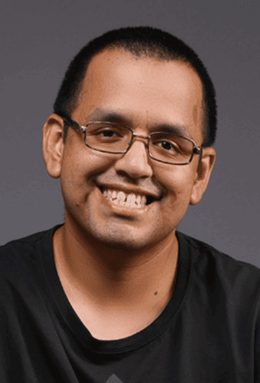 A smiling man with glasses and short dark hair wearing a black shirt against a gray background.