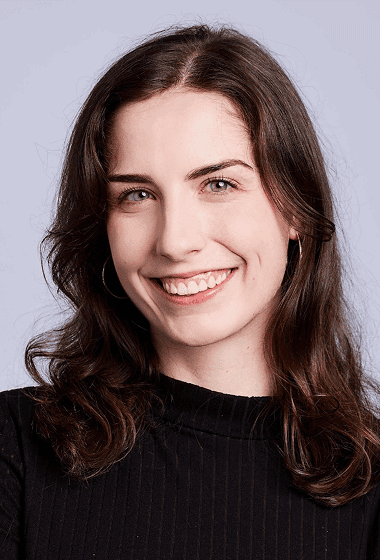 A young woman with wavy brown hair, fair skin, and a bright smile, wearing a black top and hoop earrings.