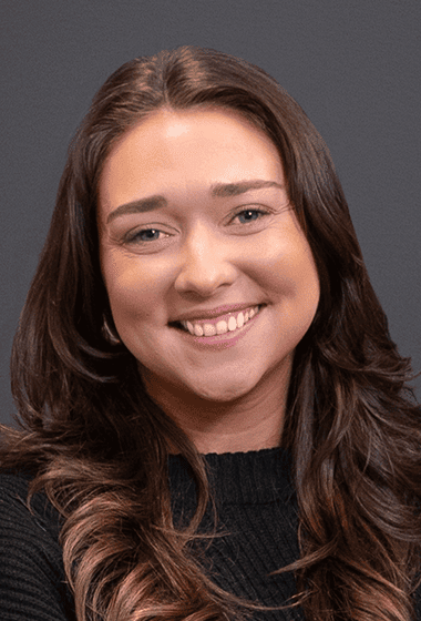 A young woman with long, wavy brown hair and a bright smile, wearing a black top against a gray background.