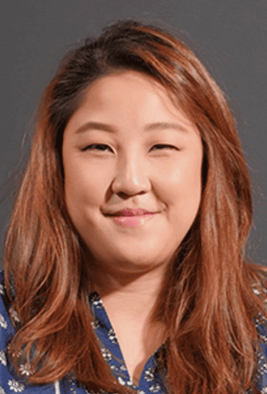 A woman with long, wavy reddish-brown hair, smiling with one eye slightly closed against a gray background.