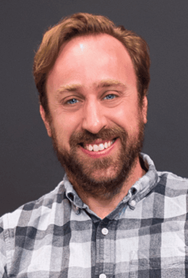 A smiling man with a beard and light brown hair, wearing a checkered shirt, against a gray background.