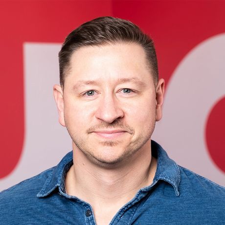 A man with short brown hair, blue eyes, and light facial hair, wearing a blue denim shirt, smiling against a red background.