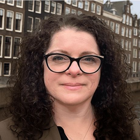 A woman with curly dark hair and glasses smiles outdoors against a background of historic brick buildings.