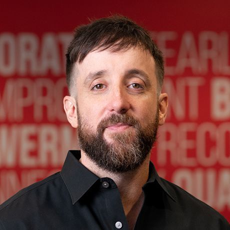 A man with short brown hair, a beard, and a black collared shirt stands in front of a red background with white text.