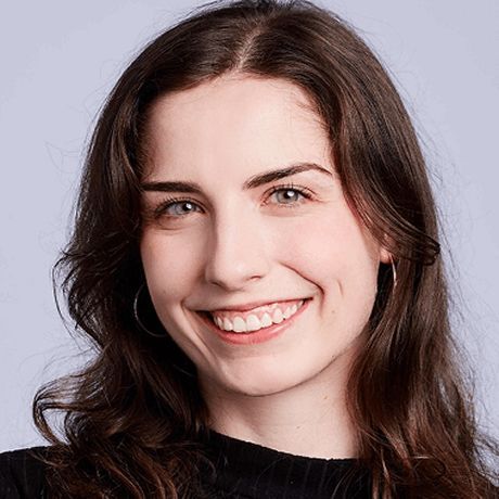 A young woman with long brown hair, blue eyes, and a bright smile, wearing hoop earrings against a light gray background.