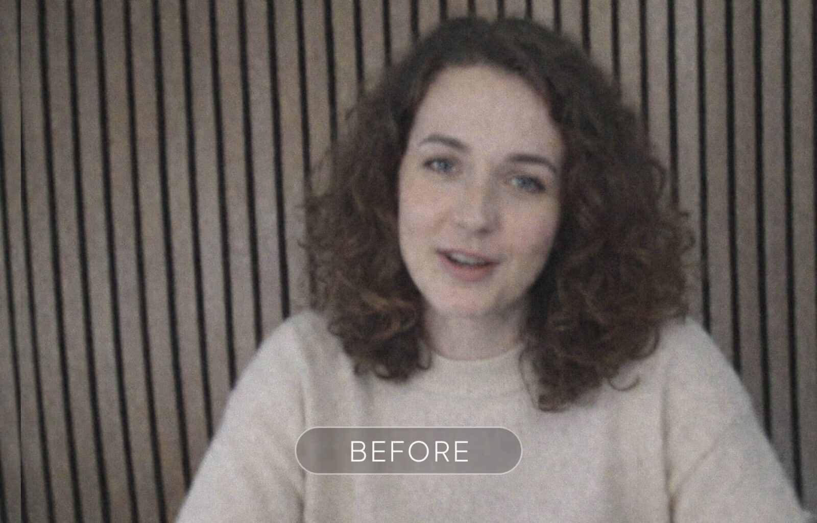 A woman with curly brown hair and blue eyes smiling in front of a striped wooden background.