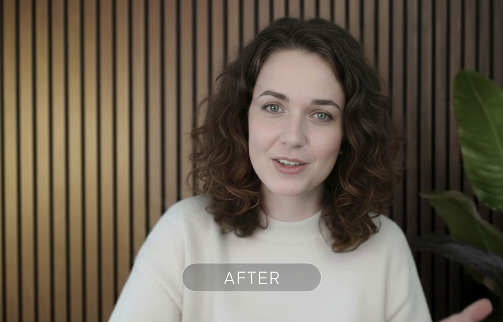 A woman with wavy brown hair and blue eyes smiling in front of a wooden slat background, wearing a white top.