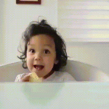 A young girl with curly hair smiling and holding a piece of bread or toast in a bright room.