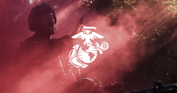 A soldier in a helmet raises his hand in front of a smoky background with a white Marine Corps emblem overlay.